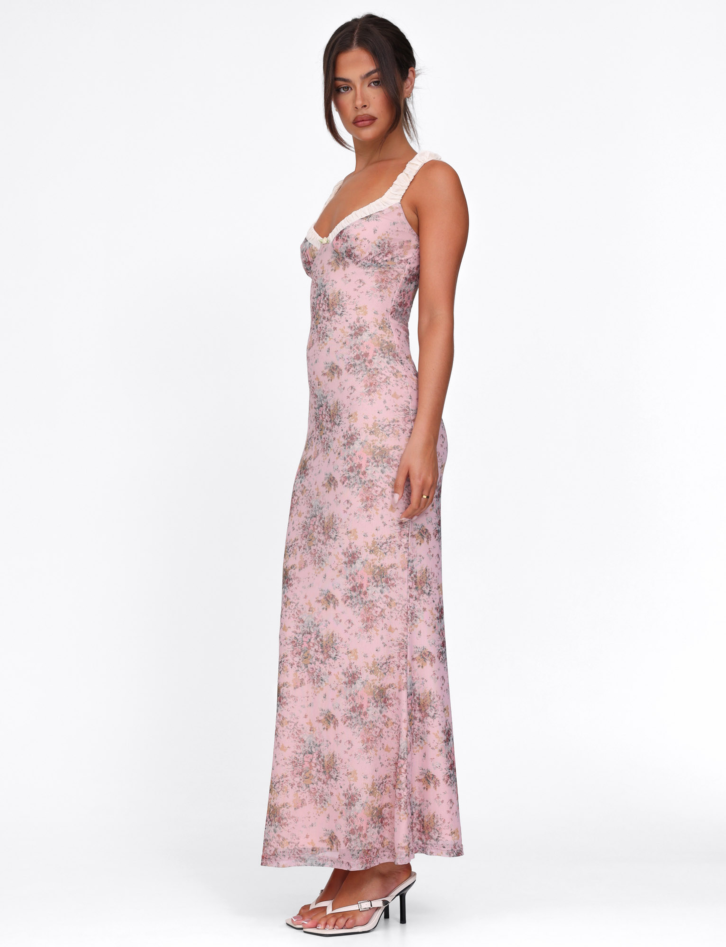 A woman poses in profile against a white background, wearing the TIGERMIST AMARAN DRESS - PINK : CAMO FLORAL, featuring a bodycon silhouette and white lace trim. She pairs it with white heeled sandals and has her hair in a loose updo.
