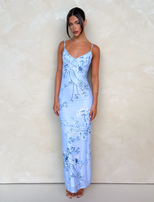 A woman with long dark hair in a loose updo wears the TIGERMIST JULIANNA DRESS in Blue Ceramic Floral, a long maxi dress with thin straps, paired with clear high heels, standing against a light beige textured wall.