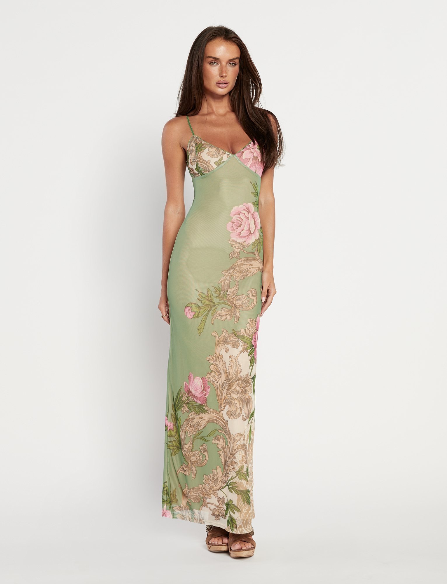 A woman with long brown hair wears the TIGERMIST JULIANNA DRESS in green Rococo Floral—a sleeveless maxi with thin straps and a pink-and-beige floral pattern—paired with brown sandals, standing against a white background.