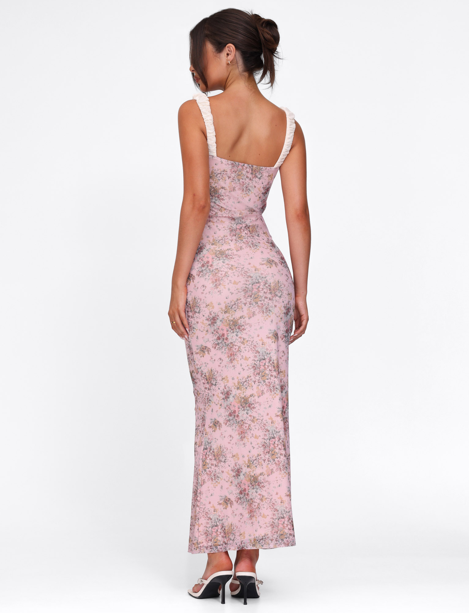 A woman with her hair tied up stands facing away against a plain white background, wearing the TIGERMIST AMARAN DRESS - PINK: CAMO FLORAL, featuring white ruffled straps, paired with black heeled sandals.