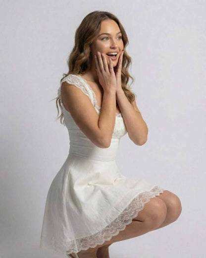 White lace mini dress with square neckline and scalloped hem, worn in a seated pose
